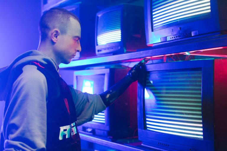 A man with a prosthetic arm interacts with vibrant vintage televisions in an industrial setting.